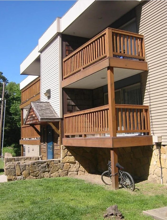 Angled front view of an apartment building with 4 balconies and an entrance in the middle. There is a bike sitting under the nearest balcony.