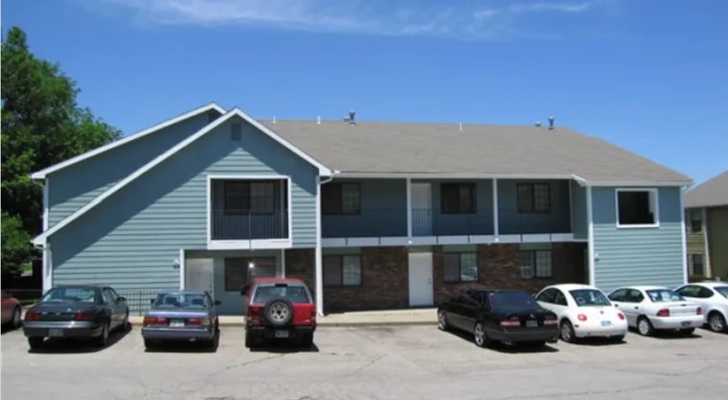 Angled front view of an apartment building with 4 balconies and an entrance in the middle. There is a bike sitting under the nearest balcony.