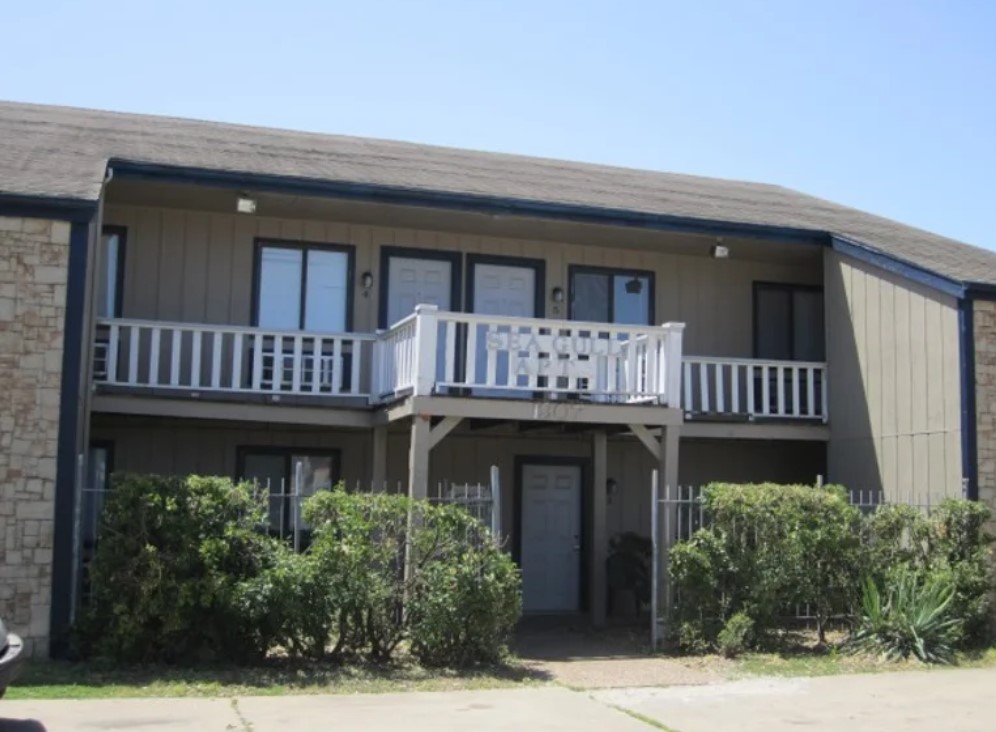 Angled front view of an apartment building with 4 balconies and an entrance in the middle. There is a bike sitting under the nearest balcony.